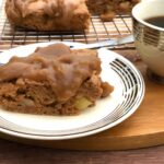 A piece of apple cake drizzled with date caramel sitting on a small plate. A cup of coffee is next to it and the rest of the cake sits behind the small plate of cake and coffee cup