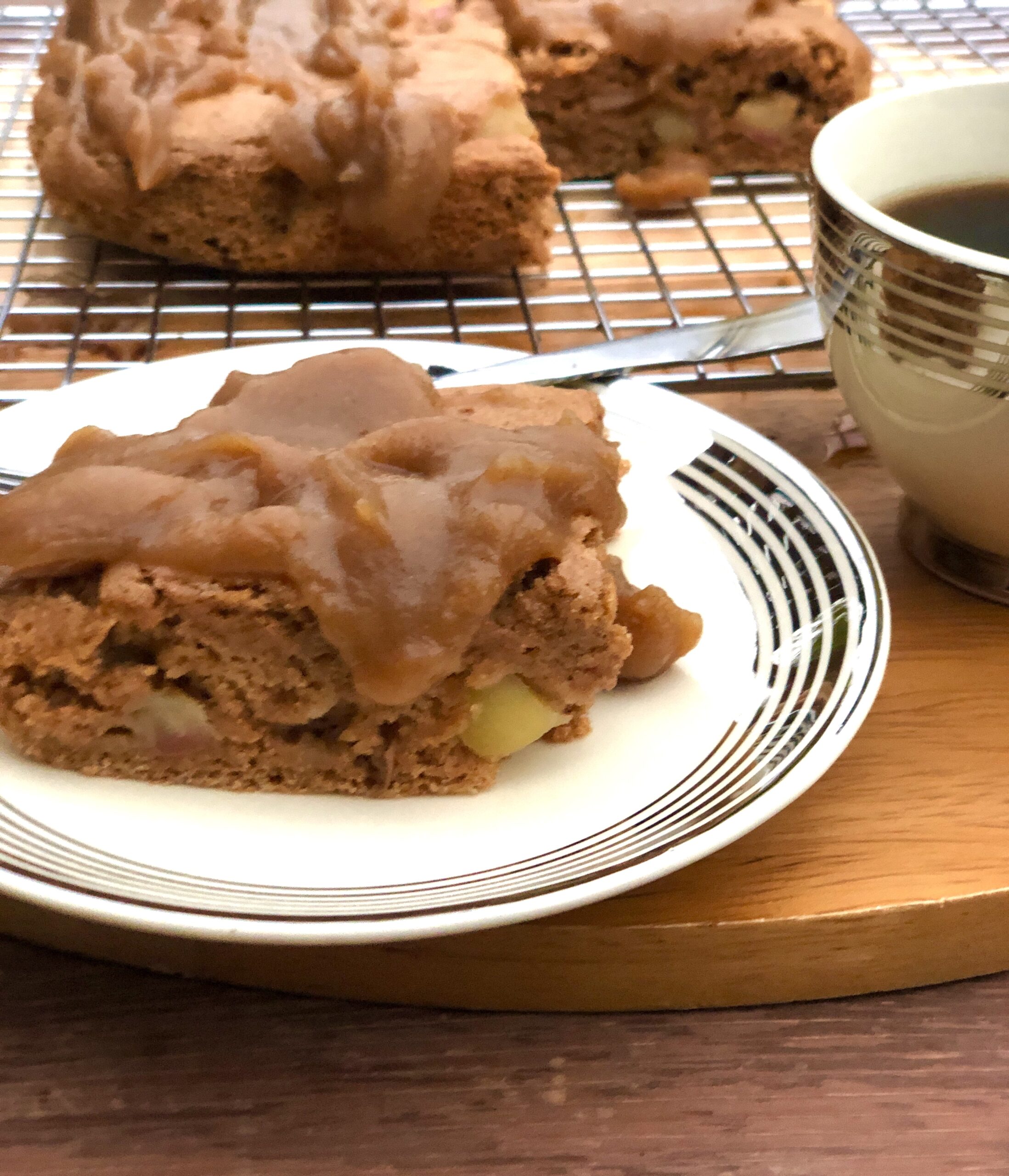 A piece of apple cake drizzled with date caramel sitting on a small plate. A cup of coffee is next to it and the rest of the cake sits behind the small plate of cake and coffee cup