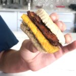 A hand holding a vegan breakfast sandwich. The sandwich consists of a tofu egg and shitake bacon on a biscuit, Vegan Eggy Sandwich,