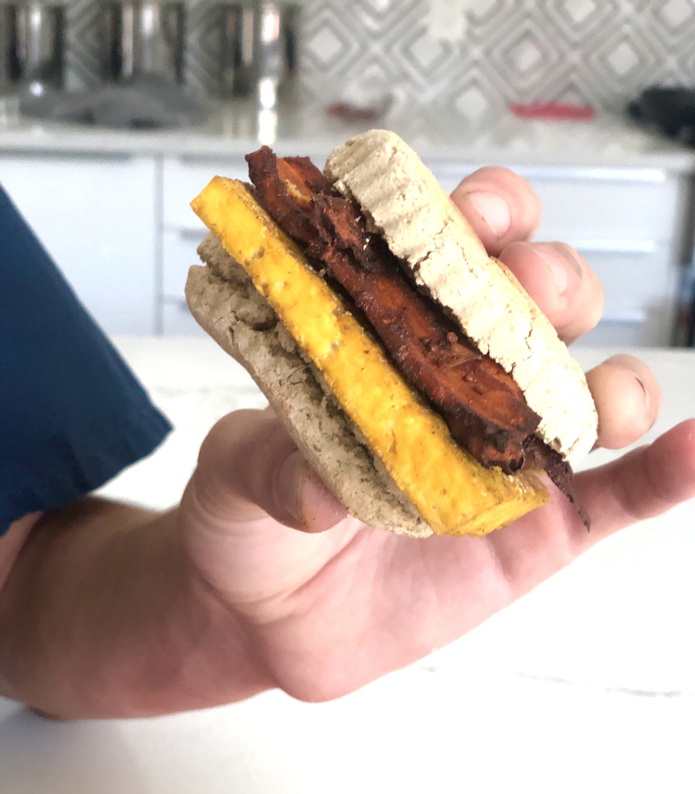 A hand holding a vegan breakfast sandwich. The sandwich consists of a tofu egg and shitake bacon on a biscuit