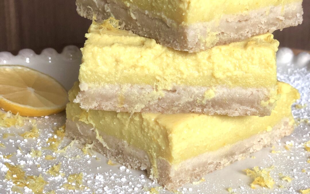 A plate with three lemon bars stacked on top of each other. The bar on top is topped with a lemon slice. The plate has lemon zest and powdered sugar sprinkled about