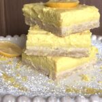 A plate with three lemon bars stacked on top of each other. The bar on top is topped with a lemon slice. The plate has lemon zest and powdered sugar sprinkled about