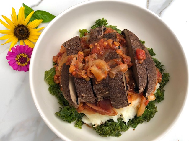 A large white bowl on a quartz countertop. There is a daisy and a Gerbera daisy in the background. The bowl contains a layer of kale, then a layer of mashed potatoes, a layer of lentil-shallot broth, and topped with sliced portobello mushrooms.
