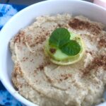 White bowl on top of a blue tie-dyed napkin. In the white bowl is a hummus spread garnished with paprika, lemon, cucumber, and mint, Fluffy Hummus,