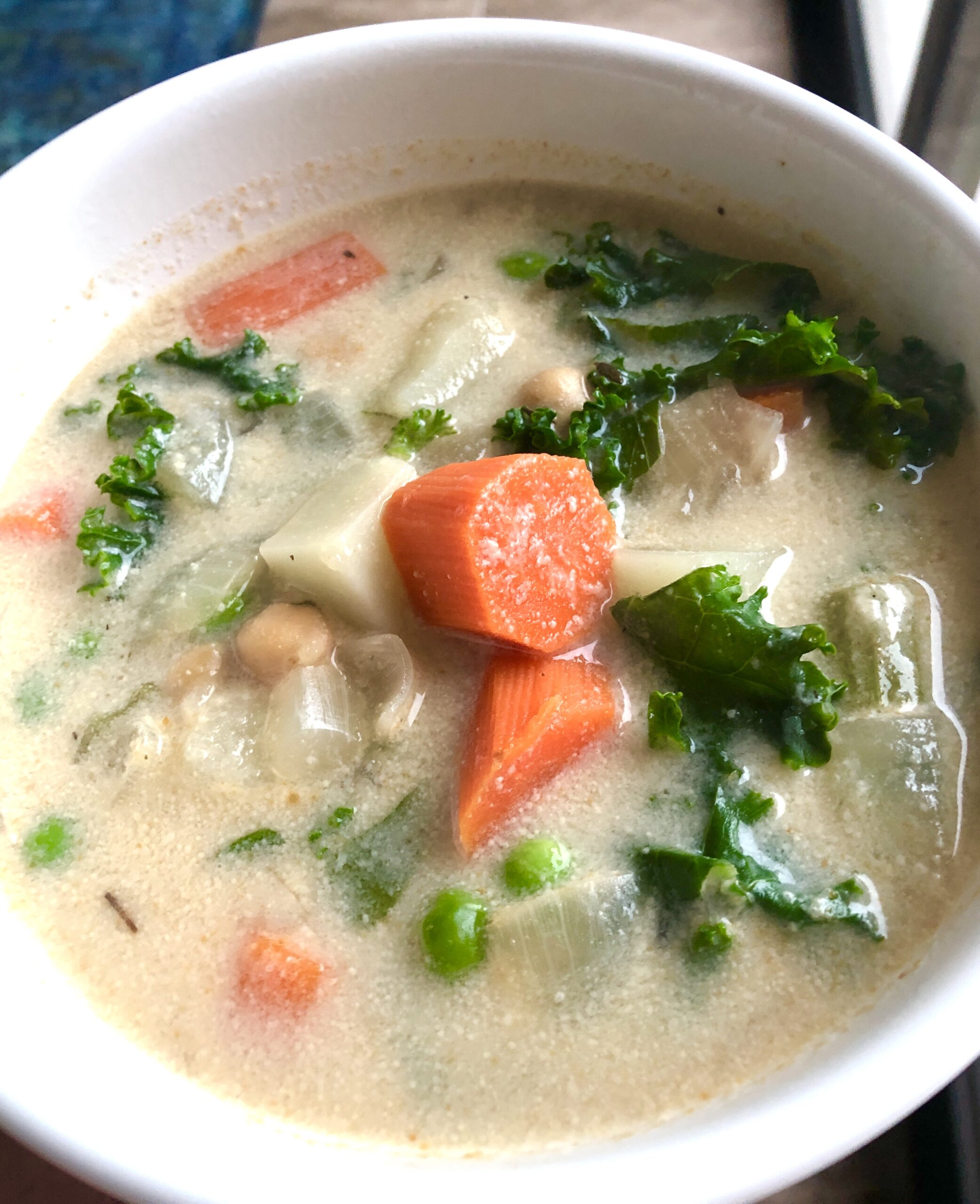 White bowl with a creamy vegetable soup. The broth is white with big chunks of carrots, potatoes, kale, and peas
