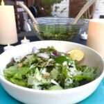 In the foreground is a white salad bowl on top of a blue plate. The salad bowl contains a salad of greens, quinoa, red onions, mint, parsley, sun-dried tomatoes, cucumbers, and pumpkin seeds with a lemon wedge in the bowl. In the background is a large serving bowl filled with salad. There are lit candles on the table