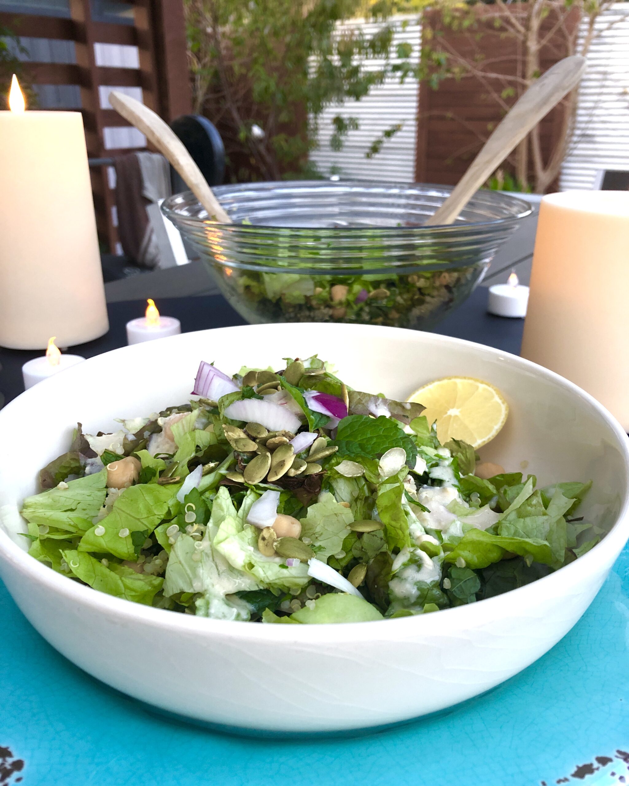In the foreground is a white salad bowl on top of a blue plate. The salad bowl contains a salad of greens, quinoa, red onions, mint, parsley, sun-dried tomatoes, cucumbers, and pumpkin seeds with a lemon wedge in the bowl. In the background is a large serving bowl filled with salad. There are lit candles on the table